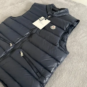 Moncler väst  - Snygg mörkblå dunväst från Moncler med quiltad design och hög krage. Västen har två dragkedjeförsedda fickor framtill och Moncler-logga på bröstet. Materialet är glansigt och lätt, perfekt för lager-på-lager. Dragkedja framtill och diskreta detaljer ger en clean look.