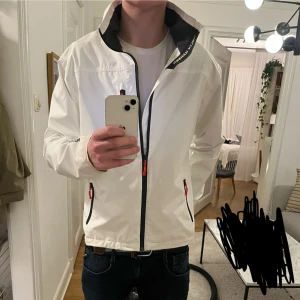 Vit seglarjacka från Helly Hansen - Snygg vit vindjacka från Helly Hansen med svart foder och detaljer i rött på dragkedjorna. Jackan har hög krage med logga, två sidofickor med dragkedja och sportig look. Perfekt för dig som gillar stilren och funktionell design. Storlek M