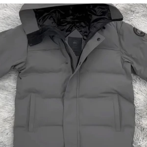 Grå dunjacka från Canada Goose - Säljer en grå dunjacka från Canada Goose med huva och svart patch på ärmen. Jackan har dragkedja och knappar framtill, två sidofickor och ett stilrent, puffigt utseende. Insidan är fodrad i svart och jackan har en clean look som funkar till streetwear.