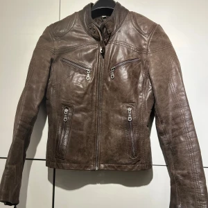 Brun skinnjacka från Celina - Snygg brun skinnjacka från Celina med coola detaljer som dragkedjor på fickor och ärmslut. Jackan har en klassisk bikerstil med ståkrage och markerade axlar. Perfekt för dig som vill ha en edgy look och gillar tidlös design.