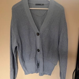 Ljusgrå stickad kofta från 157 - Snygg ljusgrå stickad kofta från 157 i storlek M. Koftan har grovstickad struktur, djup v-ringning och tre mörka knappar framtill. Långärmad och med breda muddar vid ärmslut och nederkant. Perfekt för lager-på-lager och chill stil.