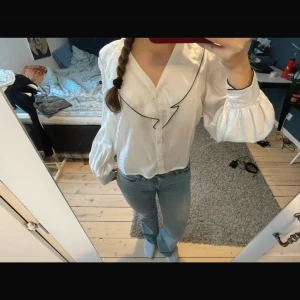 Vit blus med volangkrage XS - Supersöt vit blus med stor volangkrage och svarta kanter. Puffiga långärmar med knappar vid ärmslut och v-ringning framtill. Perfekt för dig som gillar trendiga detaljer och en feminin look.