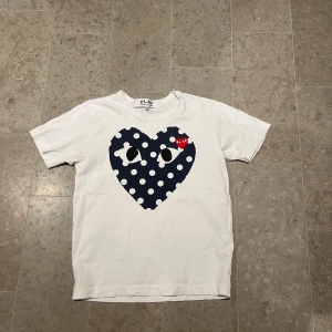 Vit Comme des Garçons Play t-shirt M - Vit t-shirt från Comme des Garçons Play i bomull med ett stort mörkblått hjärta med vita prickar och broderat rött hjärta med ögon på bröstet. Klassisk passform och korta ärmar. Perfekt statement-plagg för dig som gillar streetstyle.