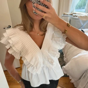 Vit blus med volang och v-ringning Zara - Supersnygg vit blus med djup v-ringning och stora plisserade volanger över axlarna och bröstet. Blusen har en loose passform och är tillverkad i bomull, vilket gör den både bekväm och luftig. Perfekt för dig som gillar statement-plagg med mycket detaljer.