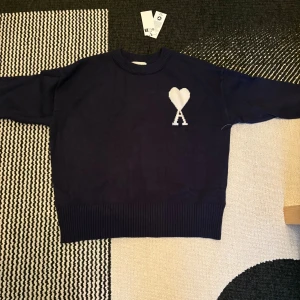 Mörkblå stickad tröja från Ami - Supersnygg mörkblå stickad tröja från Ami med vit hjärtlogga och bokstaven A på bröstet. Tröjan har rund hals, ribbade muddar och är långärmad. Perfekt för dig som gillar stilrena detaljer och vill ha något som sticker ut lite extra.