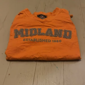 Orange Midland t-shirt med tryck - Snygg orange t-shirt från Midland med grått tryck 'MIDLAND ESTABLISHED 1880' på bröstet. T-shirten har v-ringad hals och korta ärmar. Materialet är mjuk bomull, perfekt för dig som gillar färgstarka plagg och sportig stil.