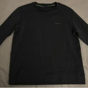 Svart sweatshirt från BOSS, storlek S - Svart långärmad sweatshirt från BOSS med diskret logga på bröstet och rund hals. Tröjan har en enkel och clean design med mjukt material och grönt band på insidan av kragen. Perfekt för dig som gillar stilrena och klassiska plagg.