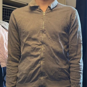 Grå kofta med dragkedja och krage - Säljer en stilren grå kofta med dragkedja framtill och klassisk krage. Tröjan har långärmad design och ribbade muddar vid nederkant och ärmslut. Perfekt för dig som gillar enkel och clean look. Materialet känns mjukt och lätt. Pris kan diskuteras!