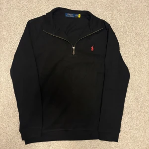 Svart half zip tröja från Polo Ralph Lauren - Svart långärmad tröja från Polo Ralph Lauren med half zip och röd broderad logga på bröstet. Tröjan har en klassisk krage och är gjord i mjuk bomull, perfekt för en chill och stilren look.