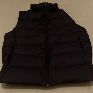 Svart dunväst från Uniqlo S - Svart dunväst från Uniqlo Ultra Light Down i storlek S. Västen är quiltad, har dragkedja framtill och praktiska fickor med dragkedja. Lätt och smidig modell som är perfekt för lager-på-lager. Klassisk, stilren look med hög krage.
