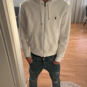 Vit hoodie från Ralph Lauren - Passade bra innan jag tvättade då ddn krympte, säljer den för att den är alldeles för liten och passar mer som S. Säljer en fräsch vit hoodie från Ralph Lauren med dragkedja och huva. Tröjan har snörning vid halsen, två fickor fram och den klassiska Polo-loggan broderad på bröstet. Perfekt för chill dagar och enkel att matcha med jeans.Knappt använd