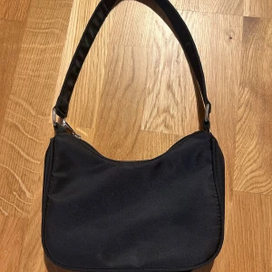 Svart axelväska från Gina Tricot - En stilren svart axelväska från Gina Tricot med enkel design och silvriga metalldetaljer. Väskan har dragkedja upptill och ett innerfack med dragkedja. Perfekt storlek för det viktigaste och lätt att bära över axeln.