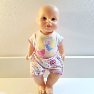 Docka med hjärtmönster och rosa kant - Ca 25 cm i längd.  Har lite tillbehör förut men de är borta! Man kunde mata den med flaskan och så kissar den på pottan.  Går att byta kläder på, tyvärr inget ombyte.   Står Newborn Baby på kläderna men vet inte märket. Minns att jag köpte den på BR.   Djur-och rökfritt hem   