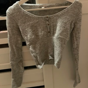 Grå stickad topp från Subdued - Säljer en grå stickad topp från Subdued med lång ärm och rundad hals. Toppen har tre dekorativa knappar framtill och är croppad i modellen. Perfekt för dig som gillar enkel och stilren design med en cozy vibe.