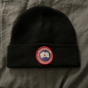 Svart mössa från Canada Goose - Svart mössa från Canada Goose med klassisk rund logopatch framtill i rött, vitt och blått. Mössan är ribbstickad och har uppvikt kant för extra stilpoäng. Perfekt för kalla dagar och enkel att matcha med din jacka.