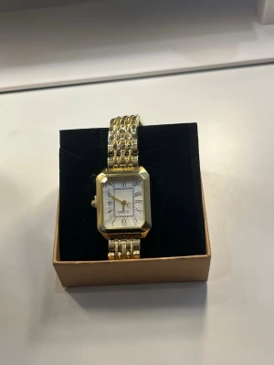 Guldfärgad fyrkantig klocka  - Stilren guldfärgad  med fyrkantigt boett och vit urtavla. Klockan har romerska siffror, guldfärgade visare och en länkad metallrem. Perfekt accessoar för dig som gillar klassisk och elegant stil.