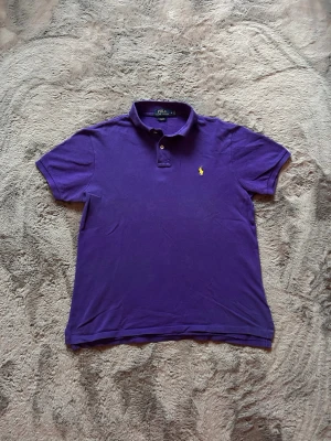 Lila pikétröja från Ralph Lauren - Klassisk lila pikétröja från Ralph Lauren med korta ärmar och krage. Tröjan har två knappar vid halsen och den ikoniska gula logotypen broderad på bröstet. Perfekt till jeans eller shorts för en avslappnad men snygg look.