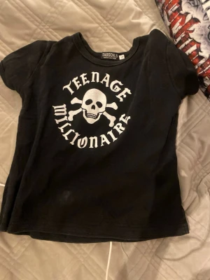 Svart Teenage Millionaire t-shirt - Står L i lappen men sitter som en S Cool svart t-shirt från Tabson med trycket 'Teenage Millionaire' och en dödskalle med korslagda ben på bröstet. Klassisk rund hals och korta ärmar. Perfekt för dig som gillar edgy streetstyle.