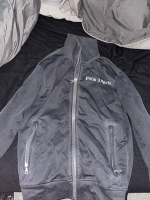 Svart ziptröja från Palm Angels - Snygg svart ziptröja från Palm Angels med hög krage och dragkedja framtill. Tröjan har två fickor med dragkedja och logga tryckt på bröstet. Perfekt för en streetwear-look och enkel att matcha med andra plagg.