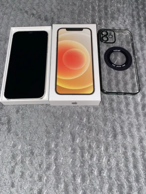 iPhone 12 med originalkartong och skal - Säljer en svart iPhone 12 med stilren design, platt skärm och tunna kanter. Telefonen kommer i originalkartong och med ett genomskinligt skal med svart MagSafe-ring. Klassisk Apple-känsla med glas på fram- och baksida samt aluminiumram.