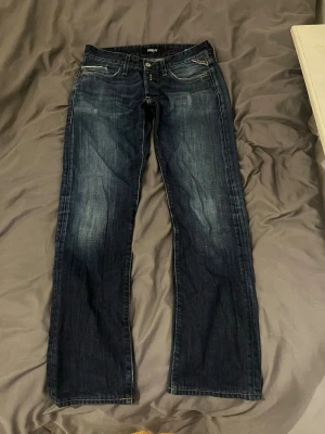 Replay blå jeans straight fit - Säljer ett par mörkblå jeans från Replay i klassisk straight fit. Jeansen har snygga slitningar framtill, fem fickor och detaljerade sömmar på bakfickorna. Tillverkade i slitstarkt denim med gul kontrastsöm och Replay-märke i midjan.