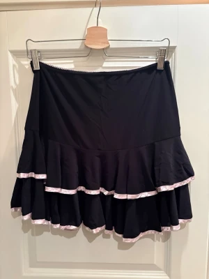 Svart volangkjol från Glitzy London - Snygg svart kortkjol från Glitzy London med dubbla volanger och ljusrosa kantdetaljer. Kjolen har en feminin och lekfull look, perfekt att styla med en enkel topp eller hoodie. Tillverkad i mjukt och följsamt material.