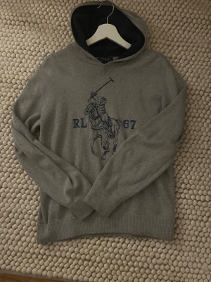 Grå hoodie från Polo Ralph Lauren - Snygg grå hoodie från Polo Ralph Lauren med klassiskt polospel-tryck och texten RL 67 i blått på bröstet. Hoodien har ribbade muddar och en mjuk huva med mörkare innerfoder. Perfekt för en chill och sportig look.