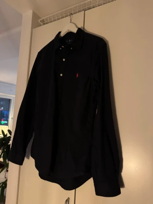 Marinblå  långärmad skjorta från Ralph Lauren - Klassisk marinblå  skjorta från Ralph Lauren med lång ärm och knappar framtill. Skjortan har den ikoniska röda logotypen broderad på bröstet och är tillverkad i mjuk bomull. Perfekt för en stilren och enkel look. Denna är dessutom i slim fit.