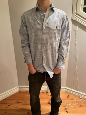 Rutig skjorta från Ralph Lauren - Klassisk rutig skjorta från Ralph Lauren i vitt och blått. Skjortan har lång ärm, knappar framtill och en liten broderad logga på bröstet. Tillverkad i bomull och har en normal passform. Perfekt för en clean och stilren look.