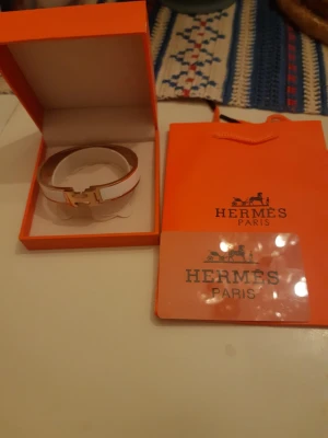 Hermès vitt armband med guldspänne - Snyggt vitt armband från Hermès med ikoniskt H-format guldspänne. Armbandet har en elegant och stilren design, levereras i original orange ask och påse. Perfekt accessoar för dig som gillar lyxiga detaljer och klassisk stil.