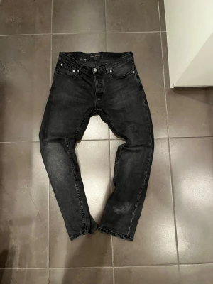 Svarta grim Tim  jeans från Nudie - Säljer ett par svarta jeans från Nudie med klassisk femficksmodell och raka ben. Jeansen har en mörk tvätt och är tillverkade i slitstark bomullsdenim. Perfekta för dig som gillar en clean och enkel look.