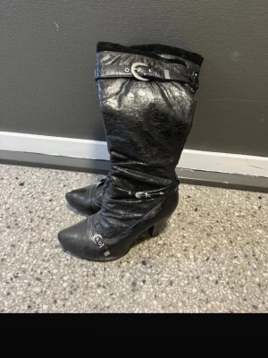 Svarta stövlar med spänne och klack - Snygga svarta stövlar i skinnimitation med spetsig tå och hög klack. Dekorativa spännen och dragkedja på sidan gör dem extra coola.  Storlek 38. London, Donna girl
