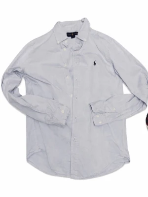 Ljusblå skjorta från Ralph Lauren - Klassisk ljusblå skjorta från Ralph Lauren med button-down krage och broderad logga på bröstet. Skjortan har långa ärmar, vita knappar och är tillverkad i mjuk bomull. Perfekt för en clean och stilren look.