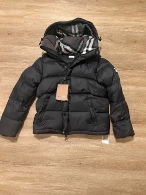 Svart pufferjacka från Burberry - Säljer en svart pufferjacka från Burberry med klassiskt rutigt foder i huvan och tydlig logga på ärmen. Jackan har dragkedja framtill, två sidofickor och en mysig, vadderad känsla. Perfekt för kalla dagar och riktigt snygg till alla outfits.