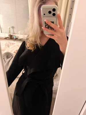 Svart omlottkappa med bälte - Snygg svart kappa med omlott-design och bälte i midjan. Jackan har lång ärm och en stilren, klassisk krage. Perfekt för dig som gillar minimalistisk stil och vill ha en jacka som funkar till många olika looks.