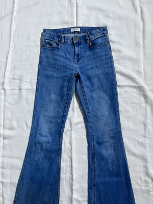 Lågmidajde blå bootcut jeans från Lindex - Säljer ett par blå bootcut jeans från Lindex med klassisk femficksdesign och bälteshällor. Jeansen har låg midja och är tillverkade i mjukt denimtyg av bomull. Snygg tvättad look och utsvängda ben för en retrovibe. Men har dock en liten fläck!! 