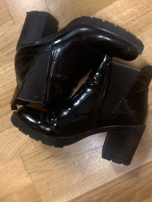 Svarta lackade stövletter med klack - Snygga svarta stövletter i glansigt lackmaterial med bred klack och rund tå. Resår på sidorna för enkel påtagning och en chunky sula som ger extra edge till outfiten. Perfekta för dig som vill ha en stilren men ändå cool look.