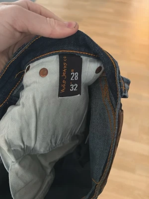 Mörkblå jeans från Nudie Jeans - Säljer ett par mörkblå jeans från Nudie Jeans med klassisk femficksmodell och kontrastsömmar. Jeansen har en smal passform och är tillverkade i slitstarkt denimtyg av bomull. Perfekta för dig som gillar stilrena och tidlösa jeans.