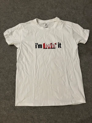 Vit t-shirt med tryck "i'm lovin' it" - Vit t-shirt med trycket "i'm lovin' it" i svart och rött på bröstet. Klassisk passform och rund halsringning. Perfekt för dig som gillar streetwear och vill ha en enkel men ändå statement t-shirt.