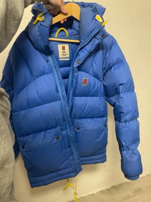 Blå Fjällräven Expedition dunjacka - Blå dunjacka från Fjällräven Expedition Series med gul snörning och detaljer. Jackan har huva, dragkedja och tryckknappar framtill samt två stora fickor. Klassisk pufferstil med Fjällräven-logga på bröstet. Perfekt för kalla vinterdagar. Herr S men passar även dam M