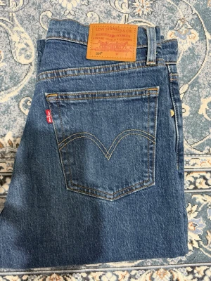 Levi's 501 blå raka jeans - Klassiska blå Levi's 501 jeans med rak passform och femficksdesign. Jeansen har den ikoniska röda Levi's-lappen på bakfickan och patch i läder bak i midjan. Tillverkade i slitstarkt bomullsjeansmaterial med snygg tvätt. Storlek: waste 72 cm, Hip 92 cm och längd 99 cm
