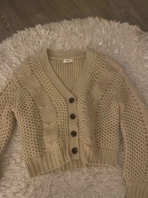 Beige kabelstickad kofta Monki - Kabelstickad beige kofta från Monki med v-ringning och fyra mörka knappar framtill. Koftan har grovstickat mönster, ribbade muddar och är croppad i modellen. Perfekt för lager på lager och en mysig look.