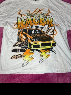Vit t-shirt med racerbil och tryck - Cool vit t-shirt med stort färgglatt tryck av en sportbil, blixtar och texten 'Kansas City Racer 42' i orange och gul. Perfekt för dig som gillar streetstyle och bilar. Skön passform och klassisk rund hals.