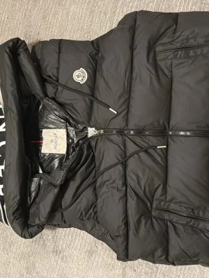 Svart dunväst från Moncler med huva - Snygg svart dunväst från Moncler med avtagbar huva och dragkedja framtill. Västen har två fickor med dragkedja och Moncler-logga på bröstet. Materialet är glansigt och fyllningen är dun, perfekt för kyliga dagar.
