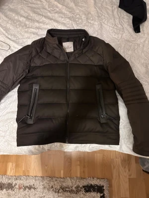 Svart dunjacka från Moncler - Snygg svart dunjacka från Moncler med quiltade partier och två dragkedjeförsedda fickor framtill. Jackan har hög krage och Moncler-logga på ärmen. Perfekt för kalla dagar och ger en stilren look.