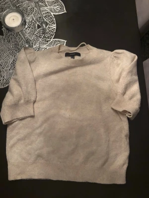 Beige stickad topp med puffärm H&M - En beige stickad topp från H&M med korta puffärmar och ribbade muddar. Tröjan har en mjuk och skön känsla, perfekt för lager på lager. Enkel och stilren design som passar till det mesta.