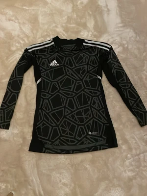 Svart långärmad Adidas tröja - Snygg svart långärmad målvaktströja från Adidas med geometriskt mönster i grått och vita detaljer på axlarna. Tillverkad i ett tekniskt material som andas, perfekt för träning eller sport. Adidas-logga på bröstet och smal passform.
