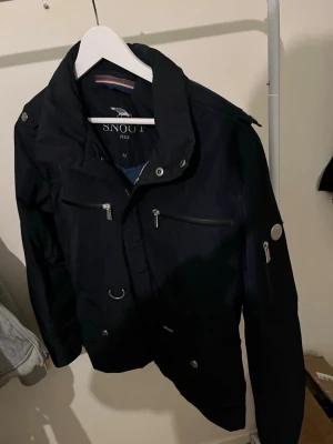 Svart Field jacka  från Snoot - Snygg svart vindjacka från Snoot med flera dragkedjeförsedda fickor och detaljer i silver. Jackan har hög krage, justerbara muddar och en modern, sportig look. Perfekt för dig som vill ha en stilren och funktionell jacka till vardags. Modellen är svår att få tag på och exklusiv. Nypris 3400. Perfekt till våren.