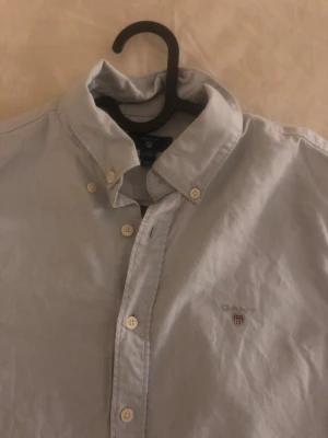 Ljusblå skjorta från Gant - Klassisk ljusblå skjorta från Gant med button-down krage och broderad logga på bröstet. Skjortan har långa ärmar, vita knappar och är tillverkad i bomull. Perfekt för en clean och stilren look.