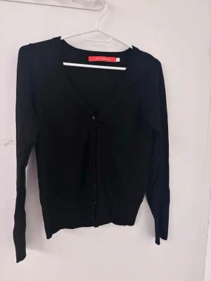 Svart kofta från Red Herring - Stilren svart kofta från Red Herring med v-ringning och knäppning framtill. Långa ärmar med dekorativa knappar vid ärmsluten ger en extra detalj. Perfekt att slänga över en topp eller klänning för en enkel men snygg look.